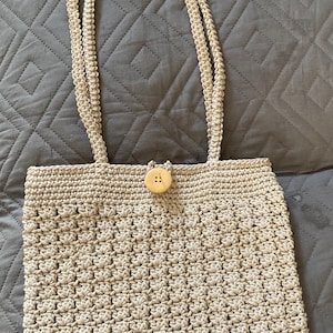 May include: A beige crocheted tote bag with a wooden button closure. The bag has a simple, classic design and is made with a thick, textured yarn.