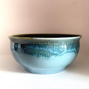 May include: A ceramic bowl featuring a smooth, light blue base that transitions into a deep, mottled teal and dark brown glaze near the rim. The bowl has a wide, rounded shape with a slightly flared edge and a glossy, reflective finish throughout.
