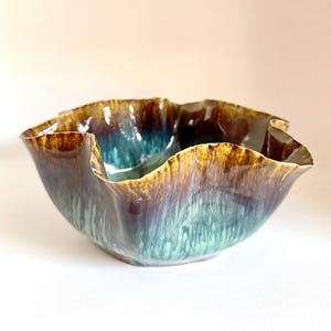 May include: A ceramic bowl with a wavy, ruffled rim featuring a glossy glaze that transitions from a deep, rich brown at the top to a vibrant teal and light blue at the base, creating a fluid, organic appearance with vertical color streaks.