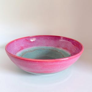 May include: A round ceramic bowl featuring a vibrant pink exterior and interior rim, transitioning into a soft teal blue centre. The glaze has a smooth, glossy finish with visible colour blending where the pink and teal meet in the basin.
