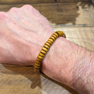 Puede incluir: Un brazalete de madera hecho de cuentas redondas apiladas. El brazalete es de color marrón claro y se lleva en la muñeca.