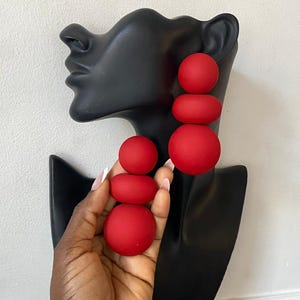 May include: Red, spherical drop earrings. The earrings are composed of three stacked, graduated-sized spheres. The earrings are displayed on a black bust against a white background. The earrings are held by a hand.