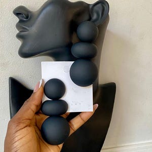 May include: Black statement earrings with a stacked, spherical design. The earrings are displayed on a black bust and held by a hand, showcasing their size and shape. The earrings have a matte finish.