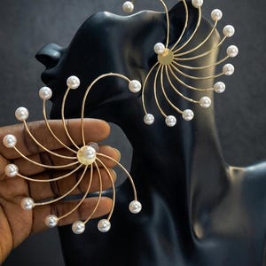 May include: A pair of gold-toned ear cuffs featuring a fan-like design with thin, curved metal wires radiating from a central circular disc. Each wire terminates in a small, round white pearl, creating a symmetrical, starburst-like appearance.
