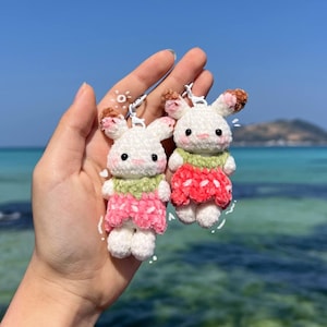 May include: Two small, white, crocheted plush rabbit keychains. Each rabbit features brown-tipped ears, black eyes, a pink nose, and a green collar. One rabbit wears a pink strawberry-patterned dress, while the other wears a red strawberry-patterned dress.