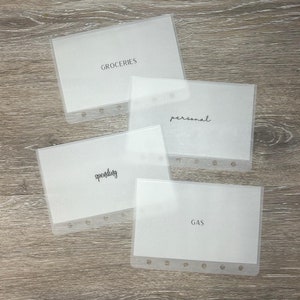 May include: Four clear plastic budget envelopes with white inserts. The envelopes are labeled "GROCERIES", "personal", "spending", and "GAS" in black script. Each envelope has a row of holes along the edge for binding. The envelopes are arranged on a wooden surface.