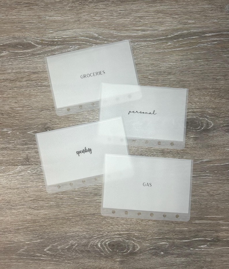 May include: Four clear plastic budget envelopes with white inserts. The envelopes are labeled "GROCERIES", "personal", "spending", and "GAS" in black script. Each envelope has a row of holes along the edge for binding. The envelopes are arranged on a wooden surface.