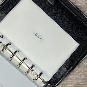May include: Black zippered binder with a silver ring binder mechanism. The binder is open, revealing a white insert labeled "GAS". The binder is made of textured black material.