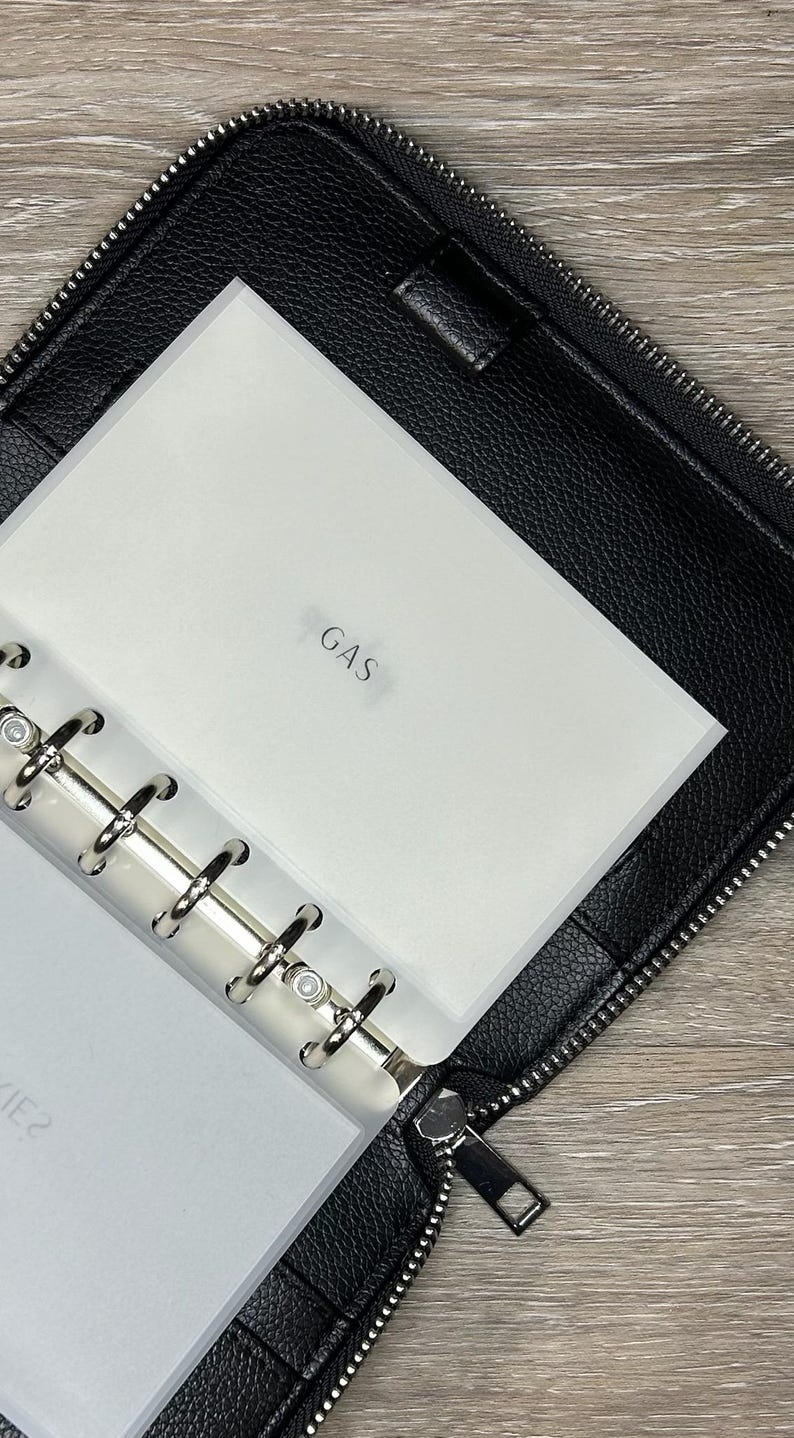 May include: Black zippered binder with a silver ring binder mechanism. The binder is open, revealing a white insert labeled "GAS". The binder is made of textured black material.
