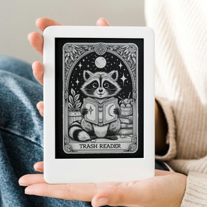 Könnte beinhalten: Ein weißer E-Reader mit einer Schwarzweißillustration eines Waschbären, der ein Buch liest. Die Illustration ist von einem dekorativen Rahmen umgeben und der Text "Trash Reader" befindet sich unter dem Bild.