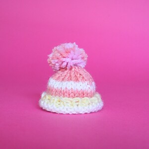 May include: A pink and white striped knitted hat with a fluffy pink pom-pom on top. The hat is on a pink background.