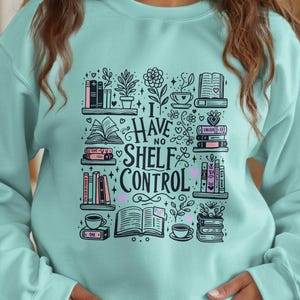 Puede incluir: Sudadera azul claro con un diseño gráfico en blanco y negro. El diseño presenta pilas de libros, tazas de café y el texto "I Have No Shelf Control". El diseño también incluye corazones y elementos florales.