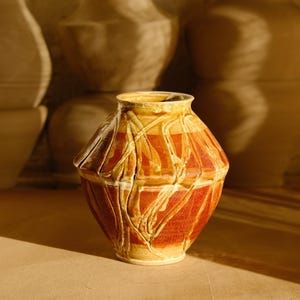 May include: A handcrafted ceramic vase with a unique design. The vase features a geometric pattern in shades of red and yellow, with a textured surface. Other similar vases are in the background. The vase is approximately 7 inches tall.