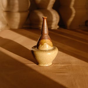 Op de afbeelding: Een handgemaakt keramisch vat met een bolvormige basis en een slanke, taps toelopende top. Het stuk heeft een warm kleurenpalet van bruin, beige en oker, en staat op een houten oppervlak. Het ontwerp suggereert een decoratief of functioneel item.