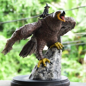 May include: A brown and gold eagle statue with a small wizard figure riding on its back. The eagle is perched on a grey rock with its wings spread out. The statue is on a black base.