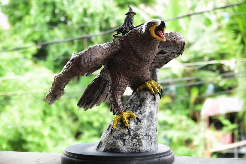 K&ouml;nnte beinhalten: Eine braune und goldene Adlerstatue mit einer kleinen Zaubererfigur, die auf ihrem R&uuml;cken reitet. Der Adler sitzt auf einem grauen Felsen mit ausgebreiteten Fl&uuml;geln. Die Statue steht auf einem schwarzen Sockel.