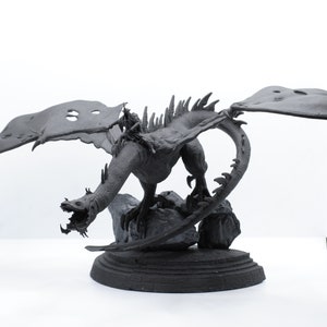 Puede incluir: Una figura de drag&oacute;n negro con las alas extendidas, posada sobre una base rocosa. El drag&oacute;n tiene una superficie detallada y texturizada y una expresi&oacute;n feroz.