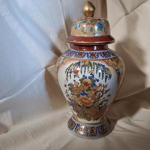May include: A large, white porcelain vase with a lid. The vase is decorated with a floral pattern in shades of blue, red, gold, and green. The vase has a gold trim around the top and bottom.