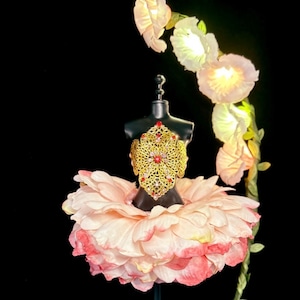 May include: A pink flower-shaped dress form with a gold and red jeweled bodice and a pink flower skirt. The dress form is on a black stand and is surrounded by white flowers.