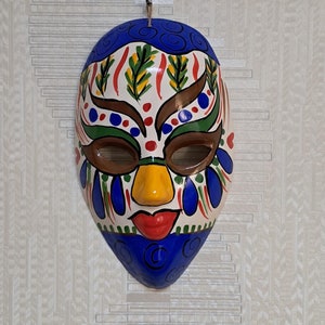May include: A ceramic mask with a blue top, yellow nose, and red lips. The mask has colourful painted details including green, red, and brown accents. It is hanging on a textured, light-coloured wall.
