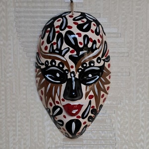 May include: A white ceramic mask with black, red, and gold painted details. The mask has a stylized human face with large eyes, a small nose, and a red mouth.
