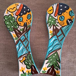 May include: Two blue ceramic spoon rests with a whimsical painted design featuring houses, trees, and birds. The houses are yellow with blue and orange details. The trees are green with white dots. The birds are yellow with black eyes.