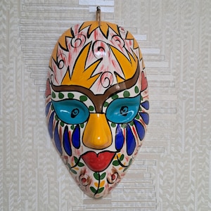 May include: A colourful ceramic mask with a yellow, red, blue and green design. The mask has a whimsical expression with large eyes and a wide mouth.