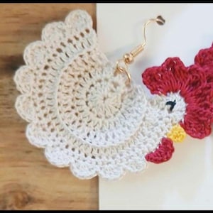 May include: A pair of handmade crochet earrings shaped like roosters. The bodies are off-white with scalloped edges, featuring bright red combs and wattles, yellow beaks, and small black embroidered eyes. Each earring hangs from a gold-tone metal hook.