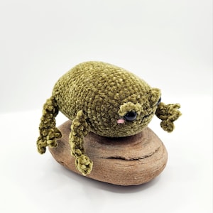 Puede incluir: Un juguete de peluche de rana de crochet verde con ojos negros y mejillas rosadas sentado en una roca marrón.