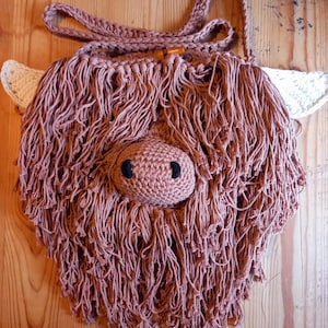 May include: A crocheted brown and white highland cow bag with a long strap. The bag has a fluffy texture and black eyes.