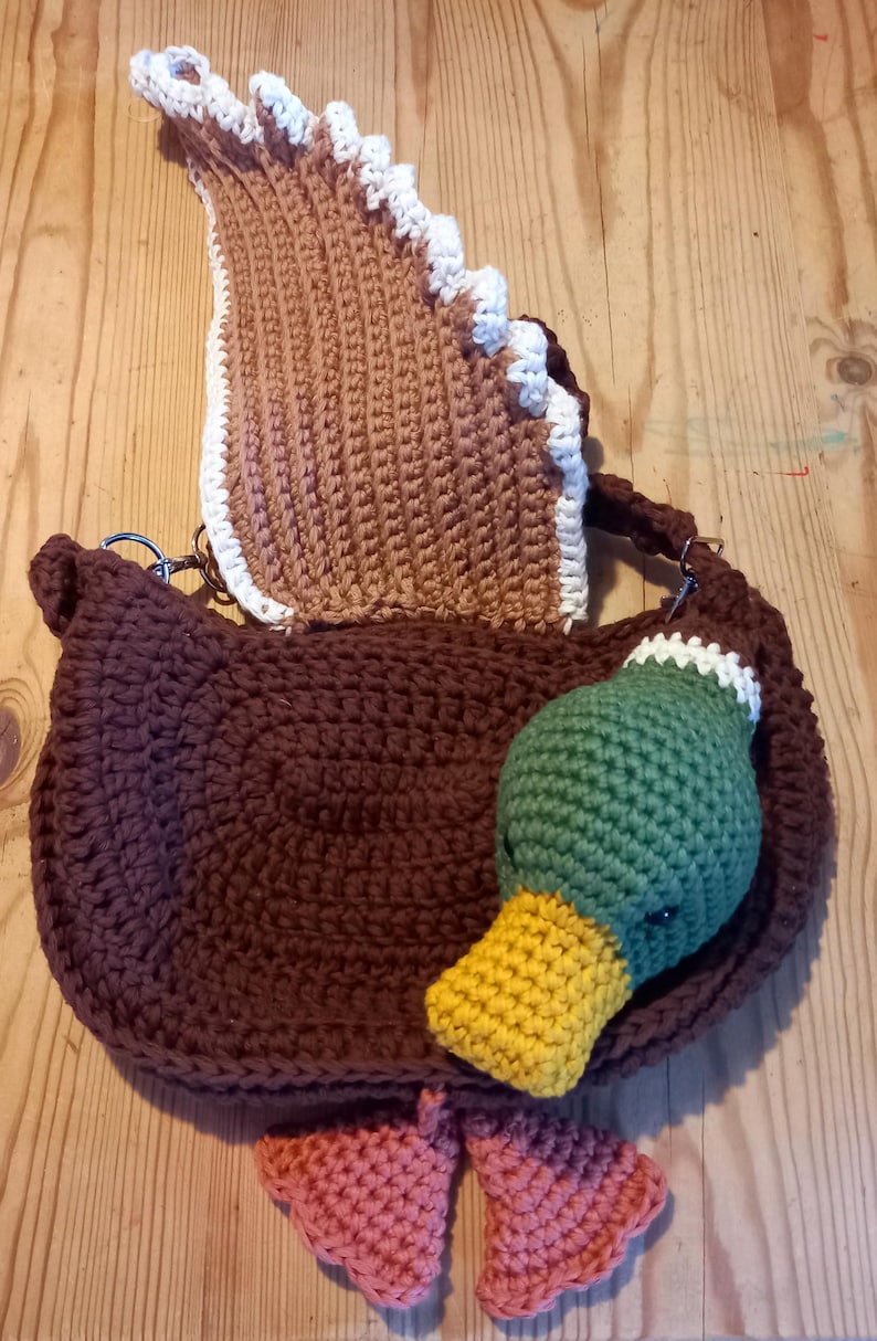 May include: A handmade crochet duck-shaped bag in brown, with a matching brown flap and a green, yellow, and white duck head. The bag has pink feet and a white trim. The bag is on a wooden surface.