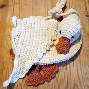 May include: A handmade crochet goose in cream and peach. The goose has a white body, head, and wings, with a peach-colored beak and feet. The wings are draped over the body. The goose is lying on a wooden surface.