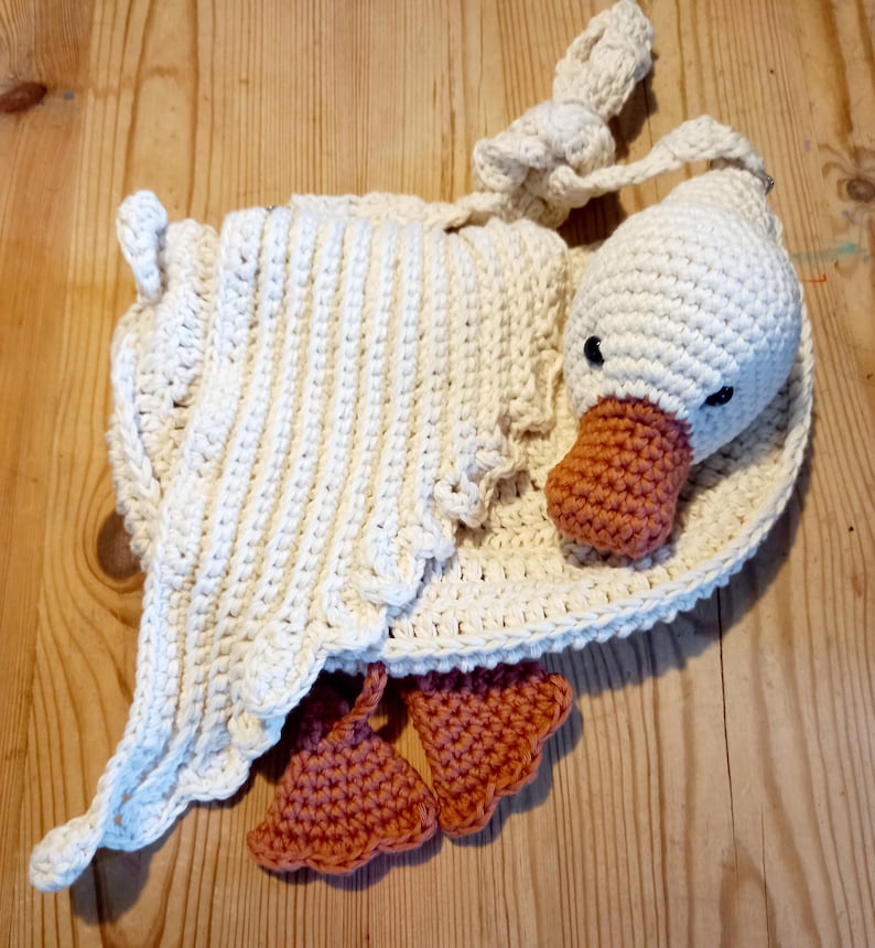 May include: A handmade crochet goose in cream and peach. The goose has a white body, head, and wings, with a peach-colored beak and feet. The wings are draped over the body. The goose is lying on a wooden surface.