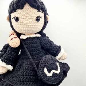 May include: A crocheted doll with black hair, a black dress, and a white crocheted collar. The doll is holding a crocheted bag.
