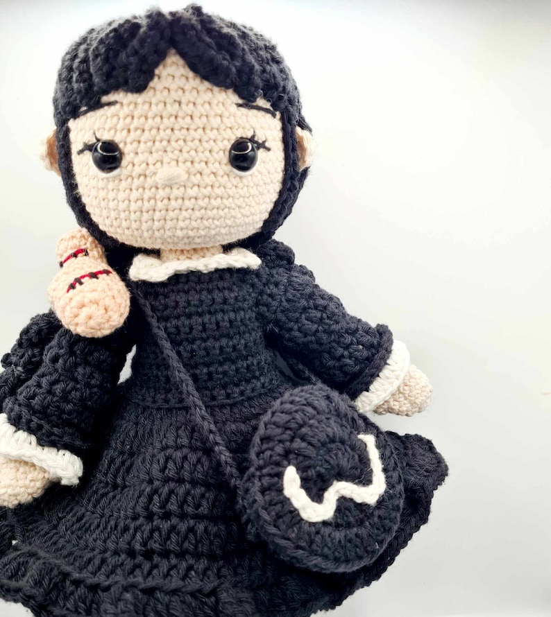 May include: A crocheted doll with black hair, a black dress, and a white crocheted collar. The doll is holding a crocheted bag.