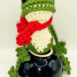 Op de afbeelding: Een handgemaakte gehaakte kikkerfiguur met een groene kop, armen en benen, een witte buik en een rode sjaal. De kikker heeft zwarte ogen en zit op een donkerblauwe keramische pot. Een grillig decoratief item.