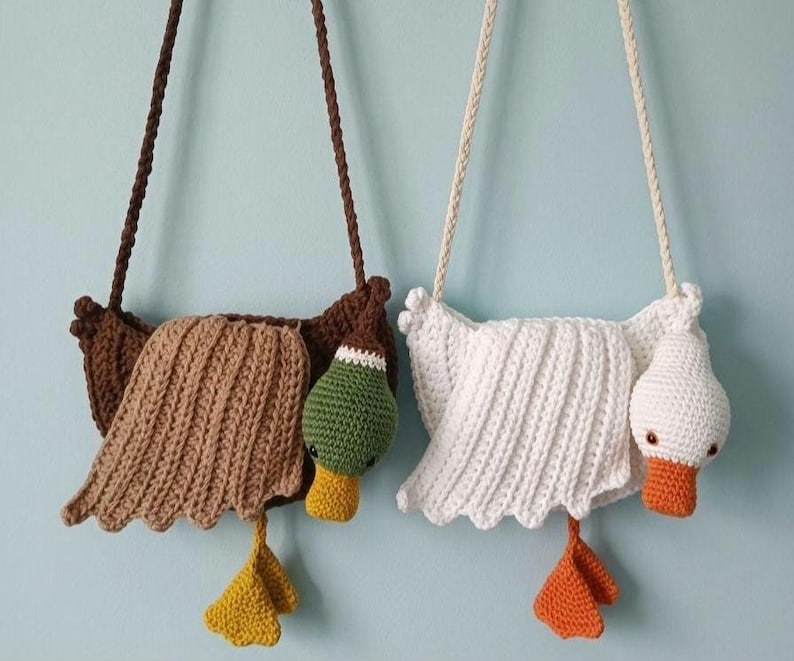 May include: Two handmade crochet purses shaped like ducks. One is brown with a green head and yellow feet, the other is white with an orange beak and feet. Both have braided straps for carrying.