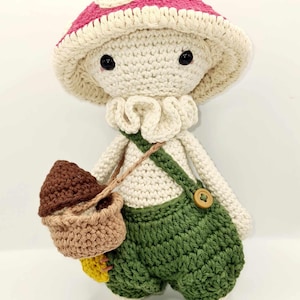May include: A crocheted mushroom doll wearing green overalls and carrying a brown acorn in a basket. The doll has a white body, pink mushroom cap, and black eyes.