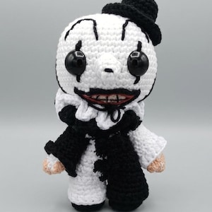 May include: A crocheted doll featuring a white head with black markings around large black button eyes, a wide mouth with visible teeth, and a small black top hat. The body is dressed in a black and white outfit with a ruffled collar and scarf.