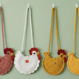 Op de afbeelding: Vier gehaakte crossbodytassen in de vorm van een kip in roze, witte, gele en oranje tinten hangen aan een lichtgroene muur. Elke tas heeft een lange riem en een knoop of decoratief element.