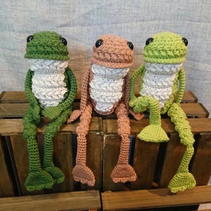 Op de afbeelding: Drie gehaakte kikkerknuffels in groen, perzik en bruin. Elke kikker heeft een getextureerd lichaam, een witte borst en zwarte knoopogen. Ze zitten op een houten krat en tonen hun lange poten en zwemvoeten. Een charmant, grillig ambacht.
