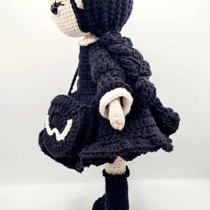 May include: A crocheted doll with black hair, a black dress with white trim, and black boots. The doll is standing on a white surface.