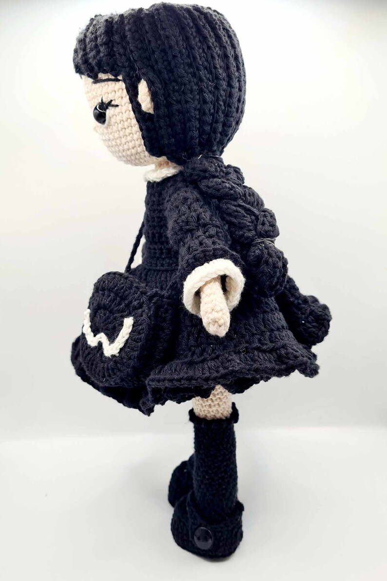May include: A crocheted doll with black hair, a black dress with white trim, and black boots. The doll is standing on a white surface.