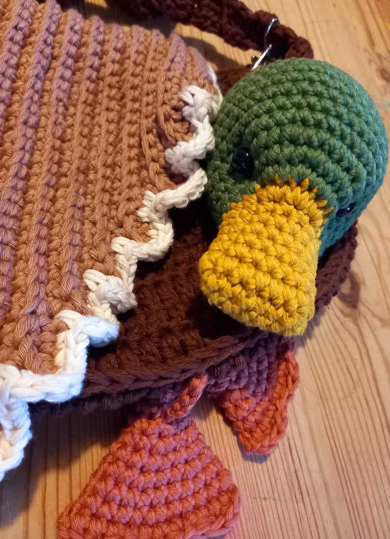 May include: A handmade crochet bag with a duck design. The bag features a brown body, a green duck head with a yellow beak, and orange feet. The bag is made with various yarn colors and textures.
