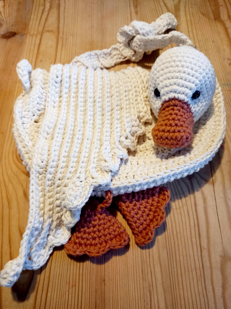 May include: Handmade crochet goose-shaped bag in cream and orange. The bag features a white goose head with black eyes and an orange beak. The body of the bag is cream-colored with a scalloped edge and orange feet.