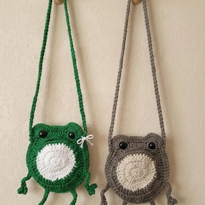 May include: Two handmade crochet frog-shaped crossbody bags. One is green with a white belly, the other is gray with a cream belly. Both have black eyes and long, braided straps. The bags are hanging on wooden pegs.