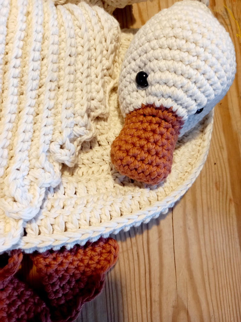 May include: A handmade crochet goose toy with a white body, head, and wings. The goose has a textured orange beak and black button eyes. The toy is sitting on a wooden surface.