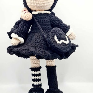 May include: A crocheted doll with black hair, a black dress with white trim, black and white striped socks, and black shoes. The doll is holding a small crocheted bag.
