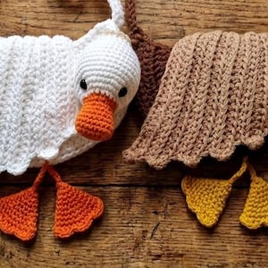 May include: Crocheted goose and duck toys. The goose is white with an orange beak and feet. The duck is green with a yellow beak and feet. Both have brown accents. The toys are handmade and sit on a wooden surface.