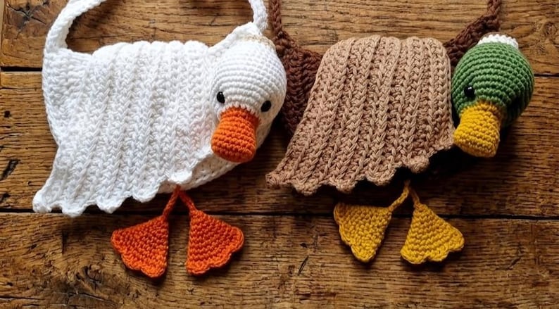 May include: Crocheted goose and duck toys. The goose is white with an orange beak and feet. The duck is green with a yellow beak and feet. Both have brown accents. The toys are handmade and sit on a wooden surface.